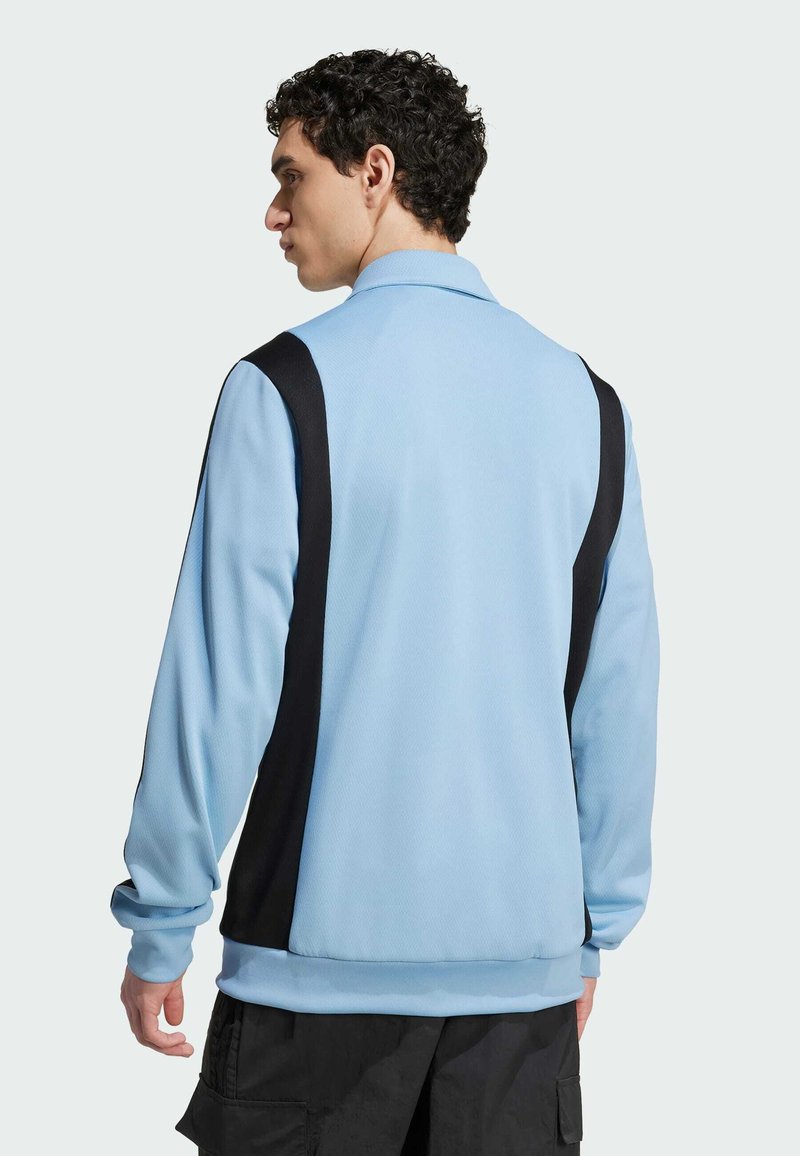 Veste pull légère bleu clair avec des panneaux latéraux noirs, en tissu texturé, à col et à manches longues. Finitions aux poignets et à l'ourlet élastiques.