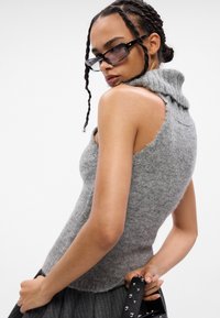 Maglione grigio senza maniche con collo a pieghe in un morbido filato testurizzato e design con spalle scoperte. La modella tiene una borsa nera con borchie.