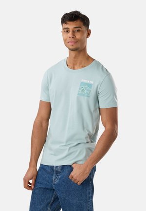 Ung mand iført en lyseblå T-shirt med motiv af en skildpadde og blå jeans, står med den ene hånd i lommen.