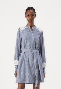 Mėlyna ir balta dryžuota medvilninė suknelė, su nėrinių akcentais ant pečių, ilgomis rankovėmis, sagčių priekine dalimi, diržo juosmeniu ir plačiu sijonu.