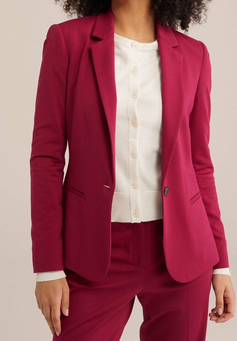 Femme portant un blazer rose foncé cintré et un pantalon assorti sur un cardigan blanc boutonné, sur un fond uni.