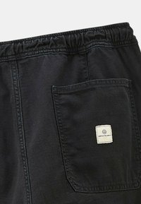Shorts noirs avec une taille élastique et une poche latérale. Présente une étiquette carrée avec le logo "Serge Blanco" sur la poche. Texture lisse.