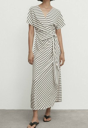 Femme portant une robe portefeuille blanche avec des rayures diagonales noires et des manches courtes, associée à des sandales noires à enfiler, debout à l'intérieur.