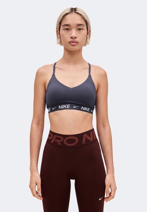 Un mannequin est debout, face à l’avant, portant un soutien-gorge de sport Nike gris foncé et un legging bordeaux sur un fond clair uni.