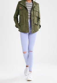 Oliwkowa kurtka typu cargo z wieloma kieszeniami, zestawiona z jasnoniebieskimi, podartymi obcisłymi jeansami oraz białymi tenisówkami. Pod spodem koszulka w paski.