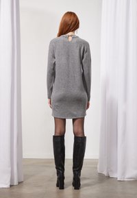 Robe grise en tricot avec manches longues et détail dos en forme de goutte, associée à des bottes noires montantes jusqu'au genou et des collants transparents. Texture lisse sur toute la surface.