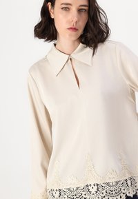 Blusa crema con colletto a punta, scollatura a occhio di bue e orlo in pizzo nero ricamato. Maniche lunghe e texture liscia su tutta la superficie.