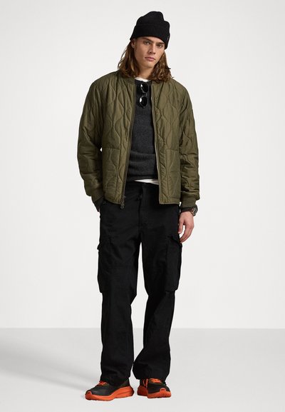 Chaqueta bomber acolchada de color verde oliva, suéter gris, pantalones cargo negros y zapatillas negras y naranjas, con una textura estampada y un diseño funcional.