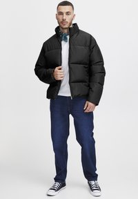 Black puffer jacket with a high collar, featuring a quilted design. Worn over a white shirt and paired with dark blue jeans and black sneakers.