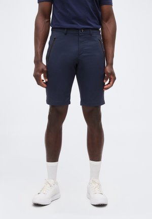 Homme portant un short bleu marine sur mesure avec poches latérales zippées, chaussettes blanches mi-mollet et baskets blanches, debout devant un fond uni.