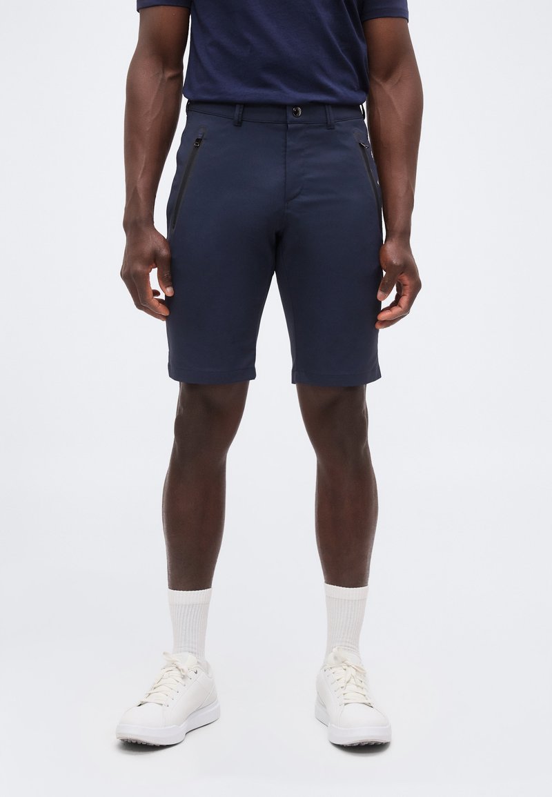 Άνδρας που φοράει σκούρο μπλε, ραμμένα στα μέτρα σορτς με φερμουάρ στα πλαϊνά τσεπάκια, λευκές κοντές κάλτσες και άσπρα αθλητικά παπούτσια, στέκεται μπροστά σε απλό φόντο.