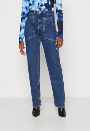 Kvinde iført højtaljede, straight-leg blå jeans med skrå forlommer, kombineret med en blå og sort tie-dye langærmet top og sorte spidse støvler.