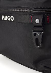 Juodas audinio krepšys su išsiskiriančiu užtrauktuku ir metaliniu užsegimu. Papuoštas raudonomis siūlėmis ir baltu "HUGO" logotipu ant audinio dirželio.