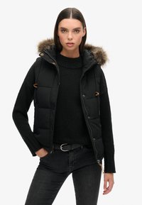 Schwarze gepolsterte Weste mit abnehmbarer, gefütterter Kapuze, Reißverschluss und Knopfleiste, kombiniert mit einem schwarzen Strickpullover und dunklen Jeans.