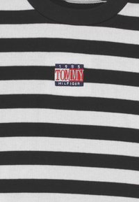 Raidallinen musta-valkoinen ribattu t-paita, jossa on pyöreä pääntie ja brodeerattu "TOMMY HILFIGER" -patch, jossa on "1985"-yksityiskohta.