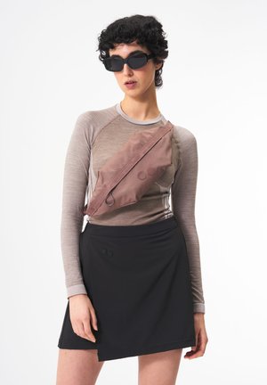 Hellbraunes, strukturiertes Oberteil mit langen Ärmeln, kombiniert mit einem schwarzen Rock. Eine braune Umhängetasche wird getragen, die zwei runde Akzente aufweist. Schwarze Sonnenbrille.