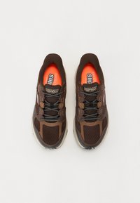 Mørkebrune sneakers med mesh og syntetiske overlay. Har reflekterende detaljer, elastiske snørebånd og en lysoransje innersåle.