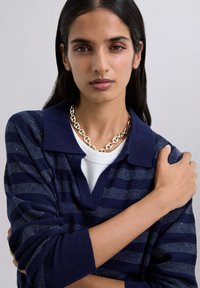Navy striped knit sweater with a polo collar, featuring textured fabric and a silver chain necklace worn around the neck.