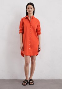 Robe chemise rouge à boutons avec col, manches retroussées et ourlet arrondi. Fabriquée en tissu lisse. Associée à des sandales noires à enfiler.