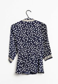 Blouse bleu marine à motif de cœurs blancs, manches longues, ourlet à basque et bordure en dentelle aux poignets. Tissu lisse et léger. Suspendue à un crochet.