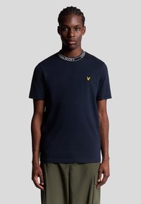 Marineblå bomulds t-shirt med rund halsudskæring og sort logo-tape detalje. Har et lille gult fugleemblem på brystet. Korte ærmer.