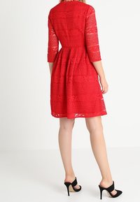 Robe en dentelle rouge avec des manches trois-quarts, des motifs complexes, un corsage ajusté et une jupe évasée. Portée avec des chaussures à talons hauts noires.