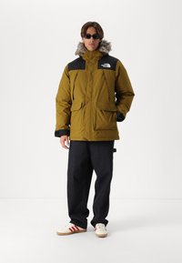 Oliven grøn parka jakke med sorte detaljer og en hætte foret med falsk pels. Har store frontlommer og en robust lynlås lukning.