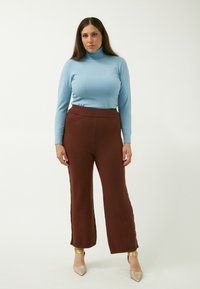 Hellblauer Rollkragenpullover mit langen Ärmeln, kombiniert mit hochgeschnittenen braunen weit geschnittenen Hosen. Das Model trägt dazu auch metallische Absatzschuhe.