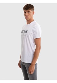 Fiatal férfi, aki fehér "BIG STAR" feliratú pólót és szürke farmert visel, oldalra fordulva áll egy egyszerű, világosszürke háttér előtt.