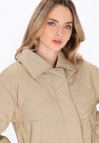 Beige quilted frakke med en stor krave, som har en glat tekstur og subtile kurvede syninger. Inkluderer en detalje med en frontlomme.