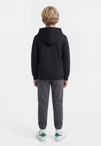 Barn med kort blondt hår iført en svart hettegenser, mørkegrå bukser, hvite sokker og hvite joggesko med grønne detaljer, stående med ryggen mot.