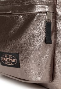 Metaliczna brązowa torba Eastpak o strukturalnym wykończeniu. Posiada kieszeń na zamek oraz czarną metkę z białym logo. Gładki i błyszczący materiał.