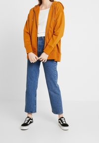 Sudadera naranja con cremallera frontal y cordones, combinada con pantalones vaqueros de tiro alto azules y zapatillas negras con suelas blancas.