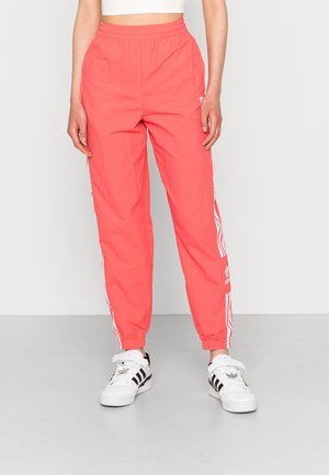 Personne portant un pantalon de survêtement Adidas corail vif avec des bandes blanches et des baskets blanches Adidas sur un fond uni clair.