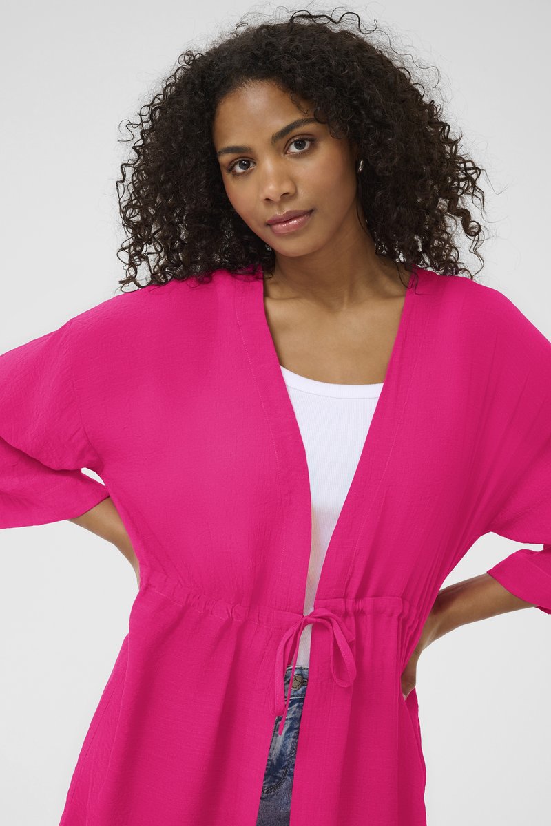 Kimono ligero de color rosa brillante con cierre de lazo y mangas anchas, que se lleva sobre una camiseta blanca. Presenta un tejido texturizado y arrugado.