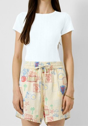 Vrouw draagt een wit topje met korte mouwen en beige korte broek met kleurrijke schelpen- en strandthema prints en een taille met trekkoord.