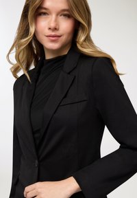 Blazer nero con design strutturato, chiusura con un bottone e vestibilità sartoriale. Presenta un rever a lancia e un dettaglio tasca discreto.