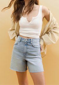 Light blue denim shorts with a high waist and frayed hem, paired with a white ribbed tank top and a beige striped oversized shirt.