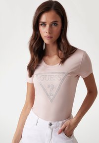 Camiseta de manga corta de color rosa claro con un logo triangular plateado de "GUESS" que incluye un signo de interrogación. Tejido suave con un diseño ajustado.