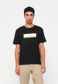 Fiatal férfi áll, fekete pólót visel, amelyen a "JACK & JONES SINCE NINETEEN NINETY" felirat, és bézs nadrágot hord egy sima háttér előtt.