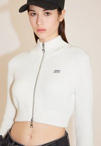 Witte geribbelde zip-up trui met hoge kraag, cropped fit, voorzien van een zilveren rits en een klein zwart logo "MISS SIXTY" op de borst.
