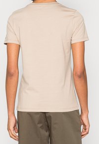 Osoba v jednoduchém béžovém tričku s krátkým rukávem a olivově zelených kalhotách, zobrazená zezadu na jednobarevném bílém pozadí.