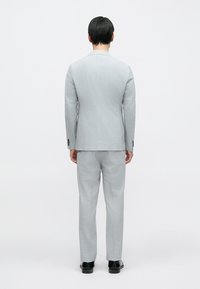 Világosszürke öltöny, testre szabott szabással, egyenes sliccel, minimális textúrával és hozzáillő nadrággal. A megjelenést fekete cipő egészíti ki.