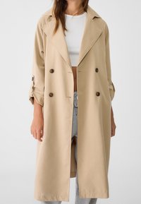 Beige double-breasted trenchcoat met opgerolde mouwen, gedragen over een witte cropped top en grijze broek met trekkoord.