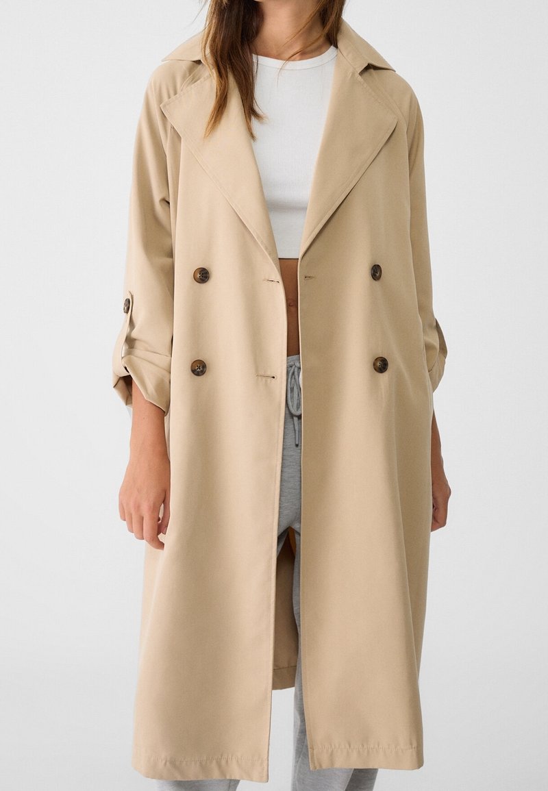 Beige double-breasted trenchcoat met opgerolde mouwen, gedragen over een witte cropped top en grijze broek met trekkoord.