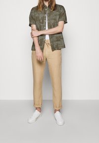 Korte mouwen, olijfgroene shirt met een patroon en knopen, gecombineerd met beige linnen broek en witte sneakers, wat een casual outfit creëert.
