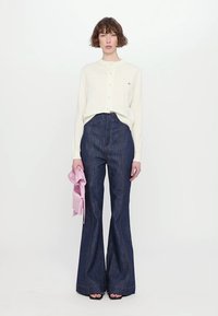 Vit, knappad cardigan över mörka denim byxor med utsvängd ben och synlig sömnad. Håller en ljusrosa accessoar. Enkla, skräddarsydda silhuetter.