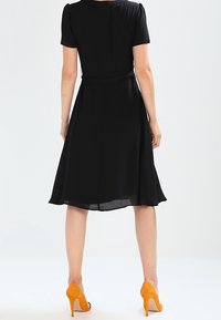 Černé šaty na krátký rukáv s upnutým pasem a rozšířenou sukní. Mají hladkou texturu a jemný lesk. Doplnené oranžovými vyššími podpatky.
