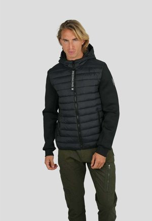 Homme blond portant une veste matelassée noire à capuche avec un détail de fermeture éclair "REDSKINS" et un pantalon cargo vert olive, debout devant un fond gris.