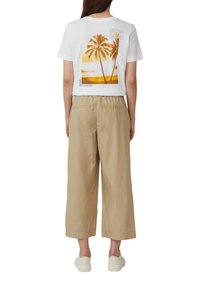 Witte T-shirt met een grafisch ontwerp van palmbomen en tekst op de achterkant, gecombineerd met beige wijde broek. Compleet met lichtgekleurde sneakers.